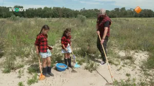 Een foto van boswachter Tim en twee kinderen in de duinen. Ze hebben allemaal een hark in hun handen en Tim harkt een stukje zand. Er staat een blauwe bak met zaden tussen de twee kinderen in. Achter hen zijn lage planten en verderop is bos te zien.