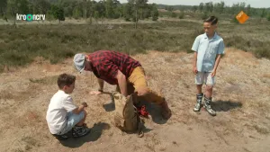 Een foto van boswachter Tim en twee kinderen in de duinen. Een kind zit op zijn hurken, Tim is bezig met naast hem op de grond te gaan zitten. Het andere kind staat aan de andere kant van Tim.