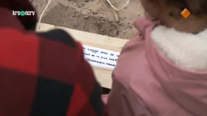 Een foto van een stuk papier met tekst dat net te zien is tussen de schouders van twee mensen. Het stuk papier zit op een plank op zandgrond.