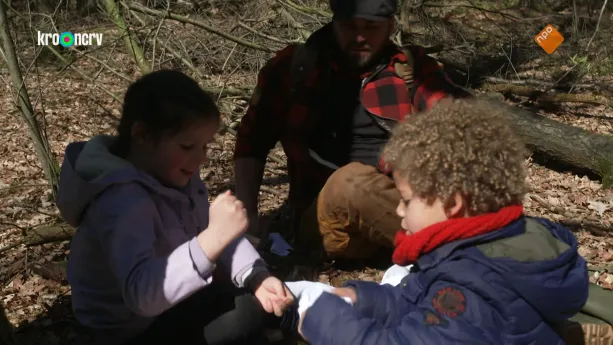 Een foto van boswachter Tim en twee kinderen. Ze zitten in de schaduw op de grond in een bos. De twee kinderen kijken naar iets wat het rechterkind in zijn handen heeft. Tim kijkt op de achtergrond toe.