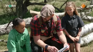 Een foto van boswachter Tim die samen met een meisje op een boomstam zitten die op de grond ligt. Er staat nog een meisje links naast boswachter Tim.