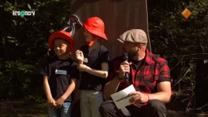 Een foto van boswachter Tim die zit en twee kinderen die naast hem staan. Hij heeft een microfoon in zijn hand en een vel papier in zijn andere hand. De twee kinderen hebben een rode regenmuts op. Ze kijken alle drie naar opzij. Achter hen zijn bomen.