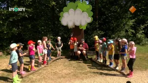 Een foto van boswachter Tim en een groep kinderen op een veldje bij een bos. De kinderen staan in twee rijen schuin links en rechts van hem. Hij zit op een stoel onder een paar witte en groene platen in de vorm van wolken.