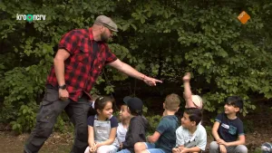 Een foto van Boswachter Tim die voor een groep kinderen staat. Hij wijst naar een kind die zijn of haar arm omhoogestoken heeft. Boswachter Tim draagt een rood met zwart geruite blouse, een beige en donkerbruin gestipte pet en een zwarte broek.