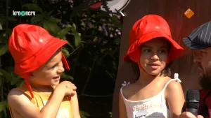 Een foto van twee kinderen met een rood hoedje op. Ze kijken naar rechts, waar Boswachter Tim zit die in een microfoon praat. De kinderen dragen zomerkleding en staan in de zon. Boswachter Tim draagt een rood met zwart geruite blouse en een zwarte pet.