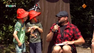 Een foto van Boswachter Tim in een rood met zwart geruite blouse aan en met een zwarte pet met grijze lijnen op. Hij praat in een microfoon in zijn hand en kijkt naar twee kinderen met een rood hoedje op die naast hem staan en ook naar hem kijken. 