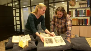 Een foto van Pascal en een mevrouw die achter een tafel staan. Op de tafel liggen zes zwarte kussens en op ieder kussen ligt een oud boek met tekst en plaatjes. Vier boeken liggen open en twee boeken rechts, die niet helemaal te zien zijn, liggen dicht.