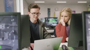 Een foto van Janouk en ethisch hacker Cas, die computersystemen test en helpt om ze beter te beveiligen. Ze zitten achter een bureau waarop allemaal beeldschermen staan en Cas zit links op een laptop te typen, waarop Janouk met aandacht meekijkt.