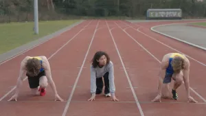 Een foto van Serah op een rode sintelbaan waar hardlopers wedstrijden lopen. Ze zit tussen twee witte lijnen op haar knie en heeft allebei haar handen voor zich op de grond voor een groene lijn om te gaan rennen, net als de twee hardlopers naast haar.