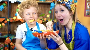 Een foto van Rosalie en Daniël. Ze hebben een rode boot die korjaal heet in Suriname, gemaakt. In de boot ligt een blauwe roeispaan, die je ook als schepje kunt gebruiken. Er liggen allemaal bananenchipjes in, waarvan Daniël er ook één in zijn mond heeft.