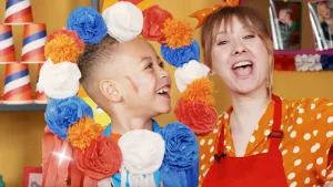 Een foto van Rosalie en D’shaedon in oranje kleding. D’shaedon heeft voor zijn hoofd een kransvan papieren rozen in rood, wit en blauw, oranje stoffen bolletjes ertussen en onderop rode, witte en blauwe papierstrookjes. Rosalie juicht met open mond.