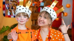 Een foto van Rosalie en Mia die een geknutselde kroon ophebben. De kroon zit met een witte stoffen band om hun hoofd en zwarte ovalen vormen zijn op de witte band te zien. Over hun hoofden heen lopen goudkleurige stroken met glinsterende rondjes erin.