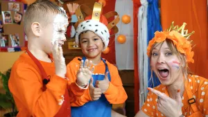Een foto van Rosalie en twee jongetjes in oranje kleding. Rosalie zit rechts en wijst naar het jongetje die witte slagroom van een taart in zijn gezicht heeft. Het andere jongetje heeft zijn duimen omhoog en een goudkleurige kroon op met een witte rand.