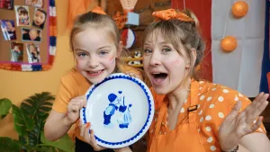 Een foto van Rosalie en Liv in oranje kleding die een Delfts blauw bord omhoog houden. Dat is een wit bord met blauwe opdruk. De opdruk is een boerenjongetje en meisje op klompen en in boerenkleding, die elkaar een kus geven met een hartje erboven.