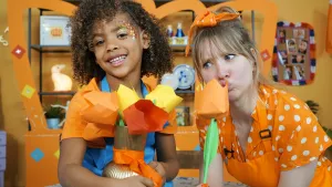 Een foto van Rosalie in oranje blouse met witte stippen, oranje schort en oranje strik in het haar. Ze ruikt aan een geknutselde oranje bloem, een tulp. Sade zit ernaast en heeft een vaas vast met andere geknutselde tulpen: oranje, geel en rood van kleur.