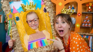 Een foto van Rosalie die met open mond wijst naar Ella. Ella zit met haar hoofd in een goudkleurige schilderijlijst. Ella's hoofd zit in het midden van het getekende kasteel in de lijst met een pluk gele vacht om haar hoofd en een kroontje op haar hoofd. 