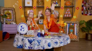 Een foto van Rosalie en een meisje die naar de camera zwaaien. Ze hebben een oranje slinger om hun nek. Voor ze op tafel staan  stroopwafels, andere koekjes en een geknutselde koning Willem-Alexander en koningin Máxima op een wit met blauw tafelkleed.