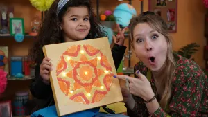 Een foto van Rosalie die naar de zandtekening in de handen van Mae naast haar wijst. Het is een goud gekleurd schildersdoek met een ronde tekening van een ster in rood, goud en oranje. In de hoeken van de ster en in het midden zit een brandend lampje.