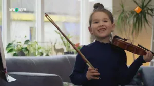 Een foto van Julia die lachend naar de camera kijkt. Julia heeft op haar linkerschouder een bruine Viool liggen. In haar rechterhand heeft ze een strijkstok vast. Op een viool maak je muziek door de strijkstok over de snaren te bewegen.