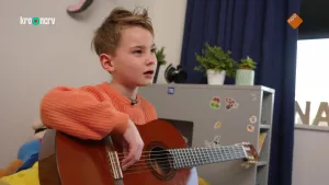 Een foto van Nathan die een donkerbruine akoestische gitaar vasthoudt en de vingers van zijn rechterhand op de snaren heeft liggen. Hij zit op zijn bed en draagt een rode trui. Op het bed achter hem ligt een knuffel van Super Mario.