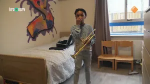 Een foto van Shahier die in zijn slaapkamer staat. Shahier heeft een trompet vast. Dat is een blaasinstrument met een ronde blaasmond, drie toetsen en een gat aan de andere kant waar de muziek uitkomt. Shahier houdt zijn rechterhand voor zijn mond. 