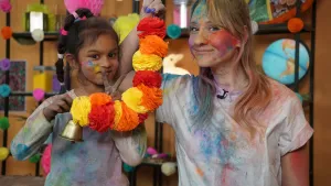 Een foto van Rosalie en Darshni die poeder van allemaal kleuren op hun gezicht, haren en shirt hebben zitten. Ze houden een slinger vast met twaalf papieren bolletjes in de kleuren rood, oranje en geel. Een goudkleurige bel hangt onder de slinger.