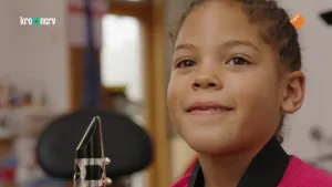 Een foto van het gezicht van Naomi en aan de linkerkant het ronde mondstuk van haar klarinet. Een klarinet is een blaasinstrument en Naomi moet in het ronde buisje blazen en haar vingers op de gaatjes leggen om muziek uit de andere kant te laten komen.