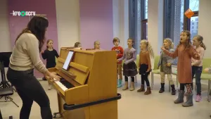 Een foto van een groep kinderen die in een cirkel staan te zingen. Aan de linkerkant staat de zanglerares achter een piano van lichtbruin hout. De zanglerares speelt op de piano en leest muzieknoten van een tablet die op een standaard op de piano staat.