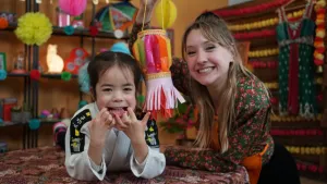 Een foto van Rosalie en Maylinn. Maylinn ligt op een tafel met uitgestoken tong en ze heeft een wit pak aan voor de zelfverdedigingssport Judo. Rosalie heeft een lantaarn vast met doorzichtig papier van allemaal kleuren waar een lichtje achter brandt.
