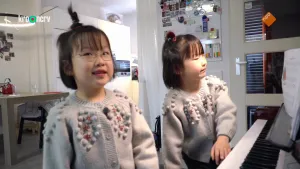 Een foto van de meisjes Kyra en Luna die achter een piano staan. Ze hebben allebei een grijs vest aan met grijze bolletjes en rode sterretjes erop. Rechts zijn de toetsen van de piano te zien en staat het boekje met de muziek erin op een standaard.