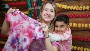 Een foto van een lachende Rosalie en Samuel. Samuel heeft zijn armen om de nek van Rosalie liggen. Rosalie houdt het t-shirt dat ze met Samuel gemaakt heeft voor de camera. Het t-shirt is wit en er staan roze en paarse cirkelvormige vlekken op.