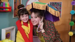 Een foto van een lachende Rosalie en Sophie. Rosalie heeft de met Sophie geknutselde paraplu vast. Op de paraplu zijn blauwe en groene vlakken zichtbaar en een paarse binnenkant. Er hangen oranje draadjes aan de zijkanten en het hengsel is lichtpaars. 