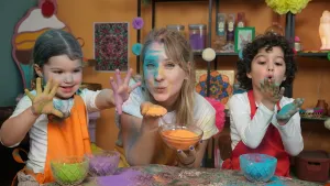 Een foto van Rosalie en twee kinderen achter een tafel. Voor de kinderen staan schaaltjes groen, paars en blauw poeder en ze houden hun handen met poeder omhoog. Rosalie heeft een schaal oranje poeder vast en wil het poeder op haar andere hand wegblazen.