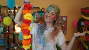 Een foto van een lachende Rosalie die een slinger van twaalf papieren bolletjes in de kleuren rood, oranje en geel vastheeft in haar rechterhand. Rosalie zit helemaal onder poeder van allemaal kleuren op haar gezicht, haren en haar witte t-shirt.  