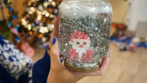 Een foto van een sneeuwbol in de handen van Rosalie. Het is een glazen potje met deksel aan de onderkant die gevuld is met water waar allemaal witte stukjes als sneeuwvlokjes inzitten. In het midden is een van kralen gemaakte kerstman te zien.