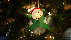 Een foto van een boom met een sneeuwpop kerstbal met een lichtje als neus en zwarte puntjes als ogen en mond. Het lichaam is een wit bolletje van stof. De pop draagt rode oorwarmers met groene bolletjes aan beide kanten en een groene strik om zijn nek.