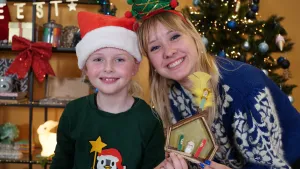 Een foto van Rosalie en een meisje. Ze hebben beiden met één hand een geknutselde kerststal vast met een puntdak. Het kindje Jezus ligt in een wit babybed tussen de rode en groene Jozef en Maria in. Op het dak zit een engel met een gele veer als vleugels.