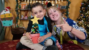 Een foto van Rosalie die naast een tafel staat en wijst naar het boompje in de handen van Ayaz die op de tafel zit. Het is een groen boompje met ronde knutselbolletjes erin en op de boom een gele ster met "Ayaz" erop. Het potje is rood met witte puntjes.