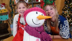 Een foto van Rosalie achter een tafel en een meisje die zit op de tafel. Vooraan staat een sneeuwpop met een rond wit hoofd, een oranje neus in de vorm van een wortel en een brede lach. De sneeuwpop draagt een paarse sjaal en een roze muts. 