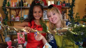 Een foto van Rosalie en een meisje achter een tak met lichtjes en takjes met groene dennenaalden eraan. Aan de tak hangen cadeautjes aan een draad met een lint als strik om de bovenkant van het cadeaupapier. Eén pakje met rood papier is helemaal te zien.