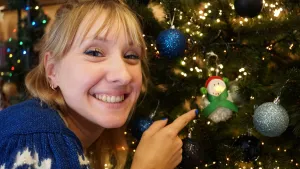 Een foto van Rosalie in een blauw met witte trui die dichtbij een kerstboom staat. Ze wijst met haar vinger naar een kerstbal in de vorm van een sneeuwpop met als neus een lichtje, op zijn hoofd een rode muts met groene bolletjes en een groene strik om.