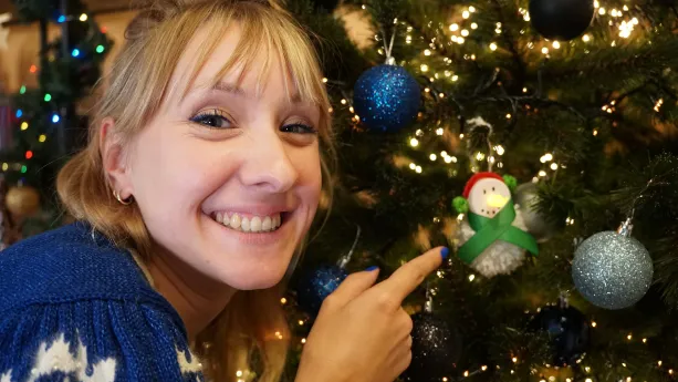 Een foto van Rosalie in een blauw met witte trui die dichtbij een kerstboom staat. Ze wijst met haar vinger naar een kerstbal in de vorm van een sneeuwpop met als neus een lichtje, op zijn hoofd een rode muts met groene bolletjes en een groene strik om.