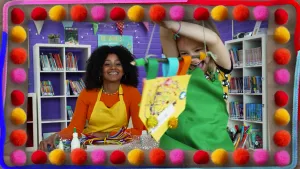 Een foto van Stephanie achter een tafel en Esmey voor de tafel. Esmey heeft een deurhanger vast aan een touwtje dat aan een takje zit met een geel papier dat met een punt nar beneden wijst eraan. Er staan bloemetjes, stipjes en glinsterende bolletjes op. 