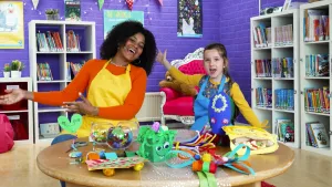 Een foto van Stephanie in gele schort en een meisje in blauwe schort die achter een tafel zitten. Ze zijn bezig "welkom bij het knutselfeest!" te roepen en houden hun armen gespreid. Op de tafel staan geknutselde cadeautjes voor één van de ouderdagen. 