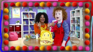 Een foto van Stephanie in gele schort die achter een tafel zit en Junne die voorop de tafel zit. Junne zit vooraan naast de snoepjesmama die hij met Stephanie gemaakt heeft. De snoepjesmama is een geel gezicht en achter de open mond liggen snoepjes.