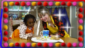 Een foto van Janayah in oranje schort en Rosalie in gele schort die naar een bord met een witte, beschilderde ontbijtmok erop kijken. Op de mok of beker staan een blauwe lucht, sterren en een halve maan. Op het bord liggen ook koekjes om de beker heen.