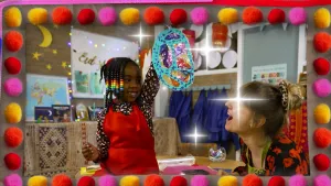 Een foto van Geneely in een rode schort die een geknutselde lantaarn van papier omhooghoudt in haar linkerhand. De lantaarn is van lichtblauw versierd papier waar vijf gedeeltes met doorzichtig vloeipapier in zitten. Rosalie kijkt er met open mond naar.