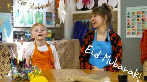 Een foto van een jongen in een oranje schort achter een tafel die naar een punt achter de camera kijkt. Rosalie zit ernaast en kijkt lachend naar de jongen. Voor Rosalie staat de tekst "Eid Mubarak!". Vertaald naar het Nederlands is dat "Gezegend Feest".