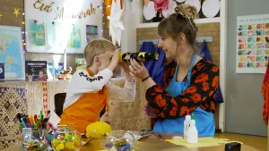 Een foto van Liam in oranje schort die door een geknutselde maankijker kijkt. De maankijker is gemaakt van een keukenrol en is zwart, met goudkleurige sterren en een maan erop. Rosalie zit ernaast in blauwe schort en heeft de maankijker middenin vast.