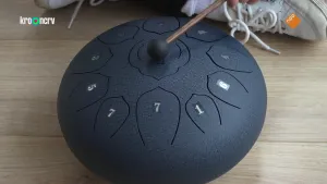 Een foto van een ronde, zwarte tongdrum en erboven een stok met zwart bolletje erop. Het is een bolle trommel met tien toetsen die half ovaal zijn en een cijfer hebben rondom en één ronde toets in het midden. Elin's witte schoenen zijn erachter te zien.